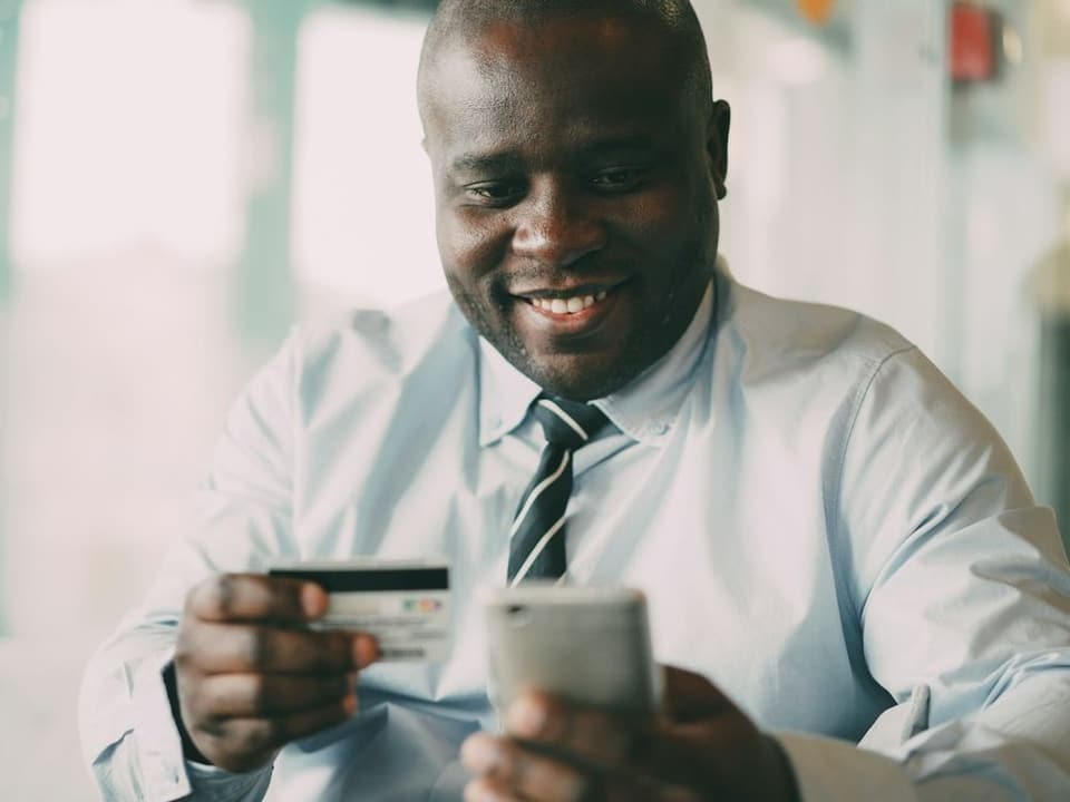 Un homme en chemise et cravate règle un achat en ligne, une carte dans une main et son téléphone dans l’autre
